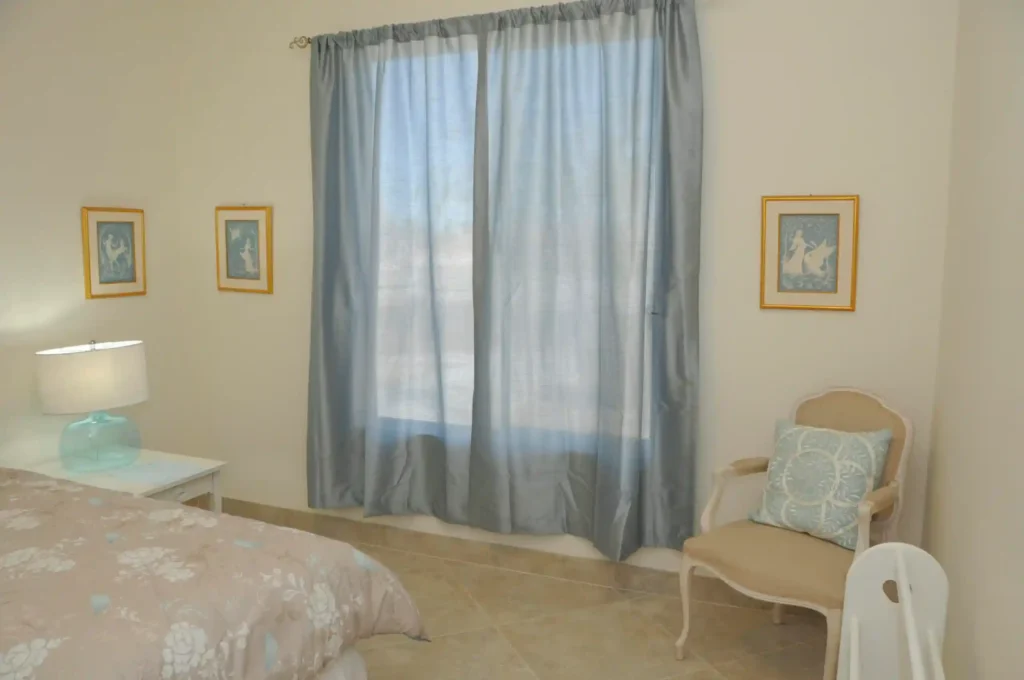 A bedroom's image in which one chair and a bed, some wall arts and a large window with a curtain you can see.
